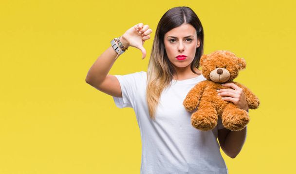 Young beautiful woman holding teddy bear plush over isolated background with angry face, negative sign showing dislike with thumbs down, rejection concept - Photo, image