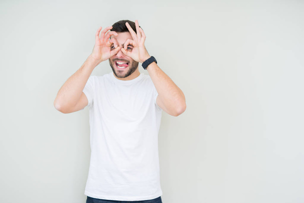 Young handsome man wearing casual white t-shirt over isolated background doing ok gesture like binoculars sticking tongue out, eyes looking through fingers. Crazy expression. - Fotoğraf, Görsel