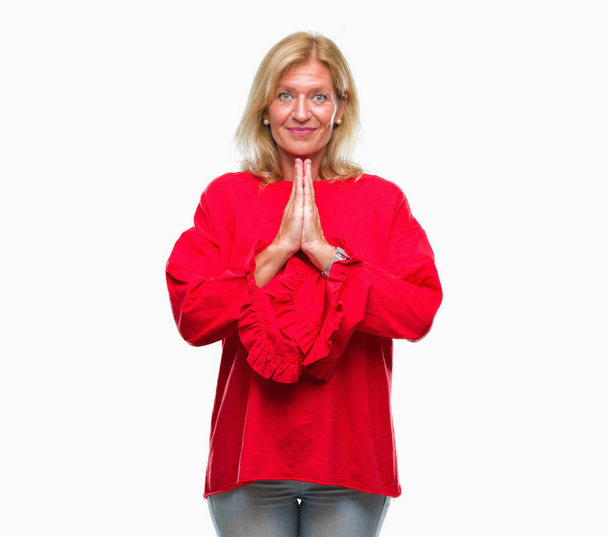 Mujer rubia de mediana edad sobre fondo aislado rezando con las manos juntas pidiendo perdón sonriendo confiado
. - Foto, Imagen