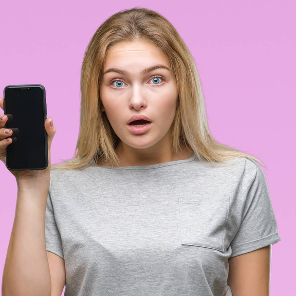 Молодая белая женщина показывает экран смартфона на изолированном фоне, напуганная в шоке с неожиданным лицом, напуганная и взволнованная выражением страха
 - Фото, изображение
