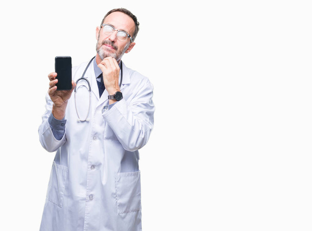 Μέση ηλικία ανώτερος γιατρός γηραιός άνδρας δείχνει smartphone οθόνη πάνω από απομονωμένες φόντο σοβαρό πρόσωπο σκέψης σχετικά με την ερώτηση, πολύ συγκεχυμένη ιδέα - Φωτογραφία, εικόνα