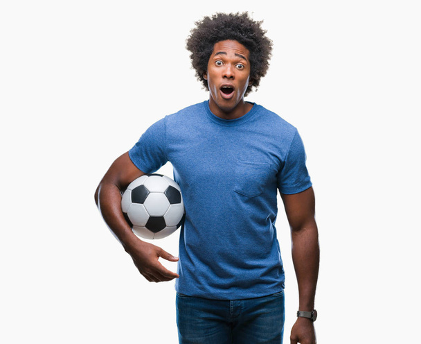 Αφρο αμερικανικό άνθρωπος που κρατά την μπάλα ποδοσφαίρου πέρα από το απομονωμένο υπόβαθρο φοβάται το σοκ με πρόσωπο έκπληξη, φοβάται και ενθουσιασμένος με φόβο έκφραση - Φωτογραφία, εικόνα