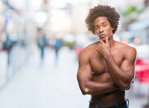 Afroamerikanischer Mann ohne Hemd, der nackte Körper vor isoliertem Hintergrund zeigt und selbstbewusst in die Kamera blickt, mit verschränkten Armen und erhobener Hand am Kinn. Positives Denken. - Foto, Bild
