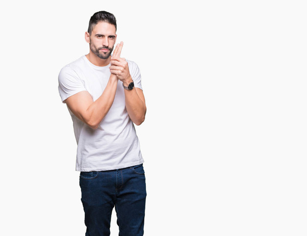 Handsome man wearing white t-shirt over white isolated background Holding symbolic gun with hand gesture, playing killing shooting weapons, angry face - Photo, image
