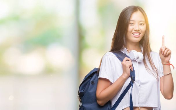 Mladá asijská žena nošení batohu a sluchátka přes samostatný pozadí překvapilo s nápad nebo otázku ukazující prst s šťastné tváře, číslo jedna - Fotografie, Obrázek