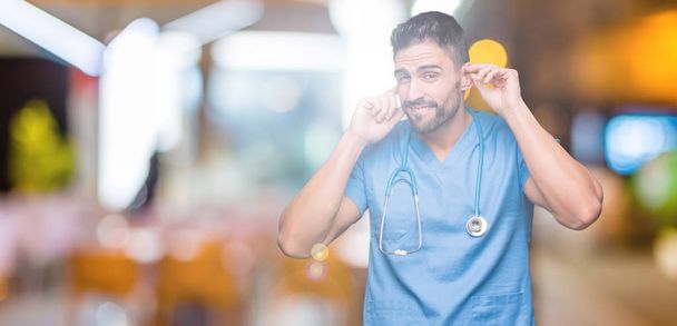 Handsome young doctor surgeon man over isolated background covering ears with fingers with annoyed expression for the noise of loud music. Deaf concept. - 写真・画像
