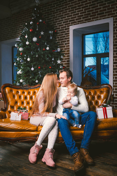thema Nieuwjaar en kerstvakantie in familiale sfeer. Stemming vieren Kaukasische jonge moeder vader en zoon 1 jaar oud zitten op een bruin lederen sofa thuis in de woonkamer in de buurt van de kerstboom. - Foto, afbeelding