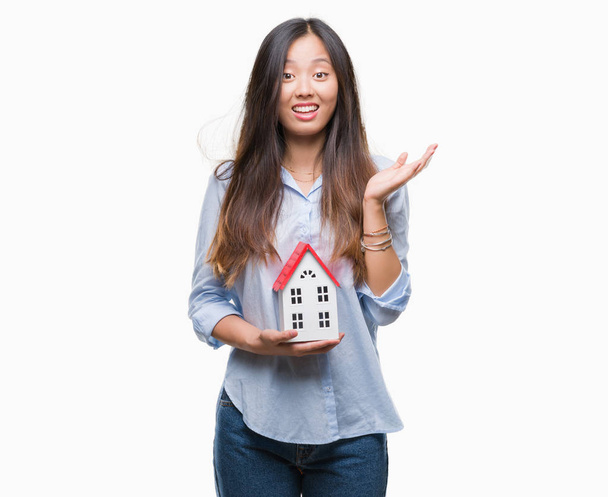 Joven mujer asiática agente de bienes raíces sosteniendo casa aislado fondo muy feliz y emocionado, ganador expresión celebrando victoria gritando con gran sonrisa y levantado las manos
 - Foto, imagen