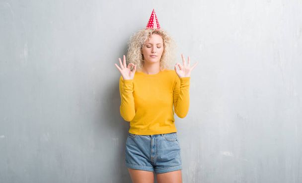 Jeune femme blonde sur un mur gris grunge portant un chapeau d'anniversaire se détendre et sourire les yeux fermés en faisant un geste de méditation avec les doigts. Concept de yoga
. - Photo, image