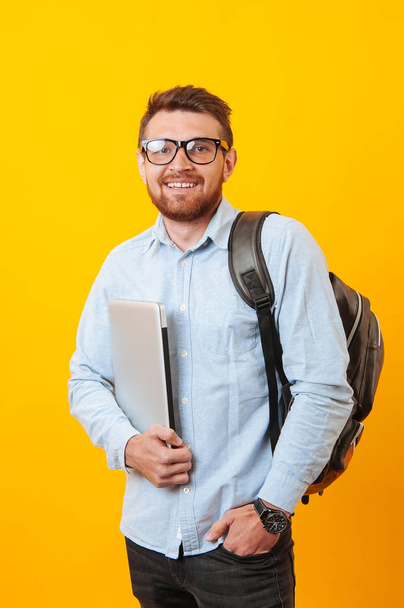 Portrait of smiling young student holding laptop and wearing backpack over yellow background - Photo, image