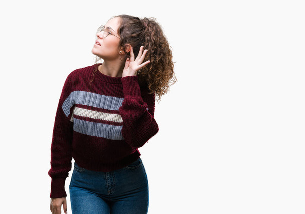 Beautiful brunette curly hair young girl wearing glasses over isolated background smiling with hand over ear listening an hearing to rumor or gossip. Deafness concept. - Photo, Image