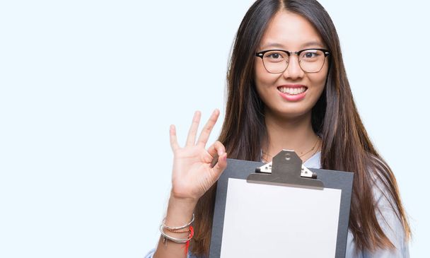 Jeune femme d'affaires asiatique tenant presse-papiers sur fond isolé faisant ok signe avec les doigts, excellent symbole
 - Photo, image