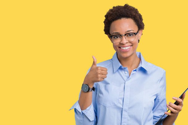 Young african american businesss woman holding clipboard over isolated background happy with big smile doing ok sign, thumb up with fingers, excellent sign - Photo, Image
