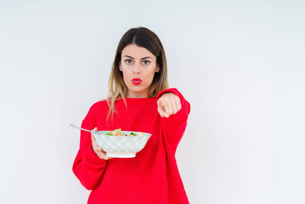 junge Frau isst frischen, gesunden Salat vor isoliertem Hintergrund und zeigt mit dem Finger in die Kamera und zu Ihnen, Handzeichen, positive und selbstbewusste Geste von vorne - Foto, Bild