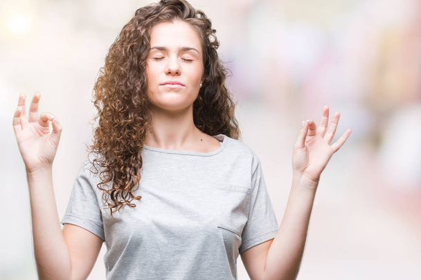 Beautiful young brunette curly hair girl wearing casual look over isolated background relax and smiling with eyes closed doing meditation gesture with fingers. Yoga concept. - 写真・画像