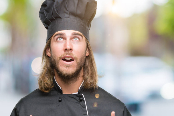 Young handsome cook man with long hair over isolated background amazed and surprised looking up and pointing with fingers and raised arms. - 写真・画像