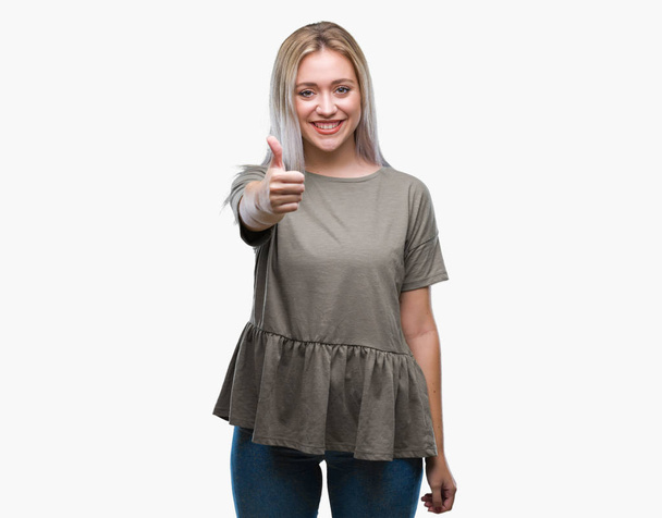 Young blonde woman over isolated background doing happy thumbs up gesture with hand. Approving expression looking at the camera with showing success. - Фото, изображение
