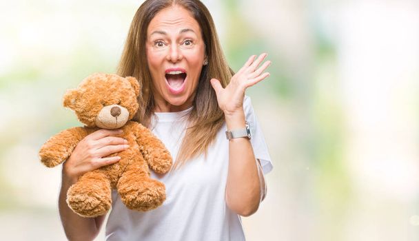 Middle age hispanic woman holding cute teddy bear plush over isolated background very happy and excited, winner expression celebrating victory screaming with big smile and raised hands - Photo, Image