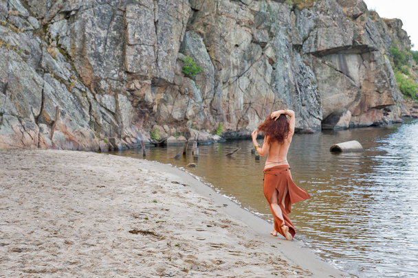 young caucasian beautiful naked Amazon woman walk turned back with raised hands on the sand river beach in front of rocks - Photo, Image