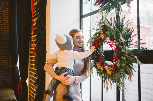 Thema Neujahr und Weihnachtsfeiertage kaukasischen Mutter hält Sohn in ihren Armen für 1 Jahr zu Hause in einem Loft-Interieur in der Nähe eines Fensters mit Weihnachten dekoriert. ein Nadelkreis wird dekoriert. - Foto, Bild