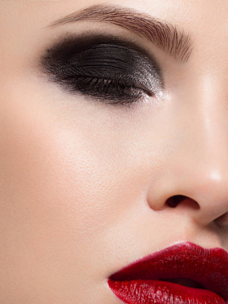 Close-up of the beauty of half a woman's face with creative fashion evening makeup. Black smoky eyes and long eyelashes, on puffy lips matte lipstick scarlet color. Well-groomed skin after spa - Photo, Image