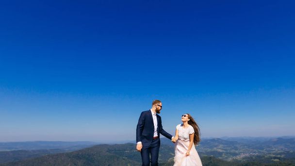 A vőlegény egy szakállas, egy elegáns ruha a menyasszony kezét a fehér ruhát, és napszemüveg tart a hegyek, a horizonton, és az ég háttere. - Fotó, kép