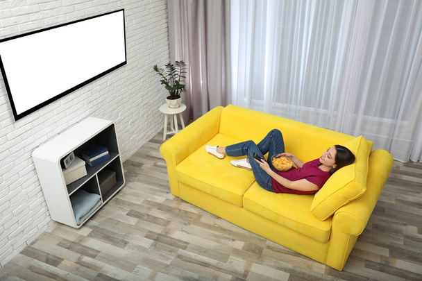 Young woman watching TV and eating chips on sofa at home, above view. Mockup for design - Foto, imagen