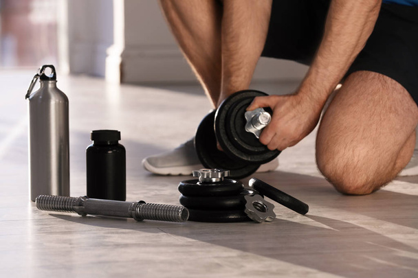 Male athlete assembling adjustable dumbbell near bottle, parts and jar of protein on floor indoors - Fotó, kép