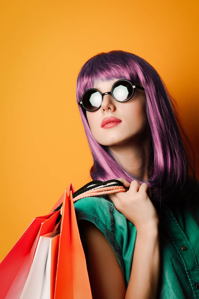 Frau mit violetten Haaren und Sonnenbrille mit Einkaufstaschen auf gelbem Hintergrund - Foto, Bild