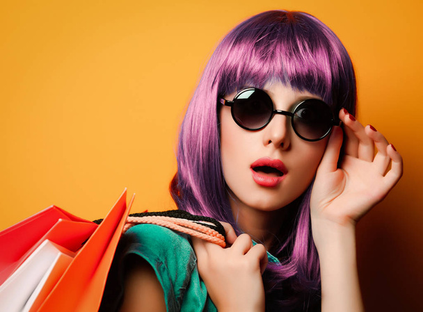 Mulher com cabelo violeta e óculos de sol com sacos de compras em fundo amarelo
 - Foto, Imagem