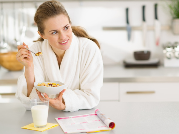 Thoughtful young woman in bathrobe eating breakfast in kitchen - 写真・画像