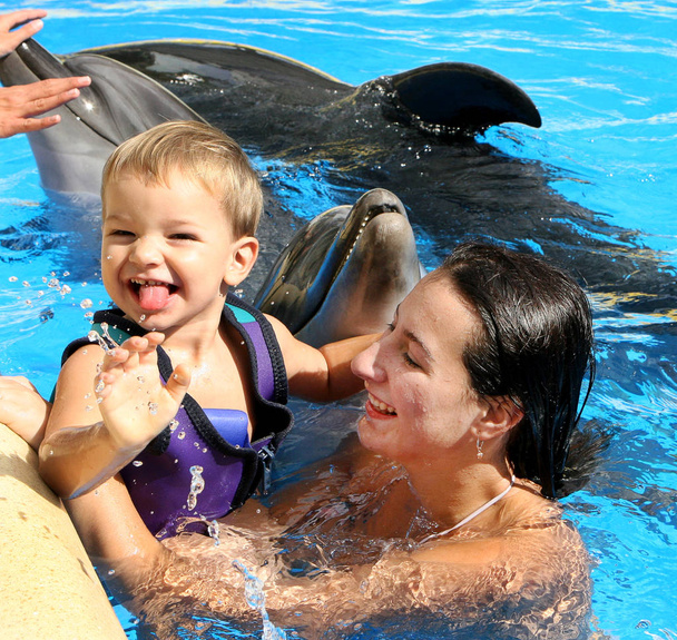 glücklich schöne junge Frau mit einem kleinen Kind lacht und schwimmt mit Delfinen in blauem Schwimmbad an einem klaren, sonnigen Tag - Foto, Bild