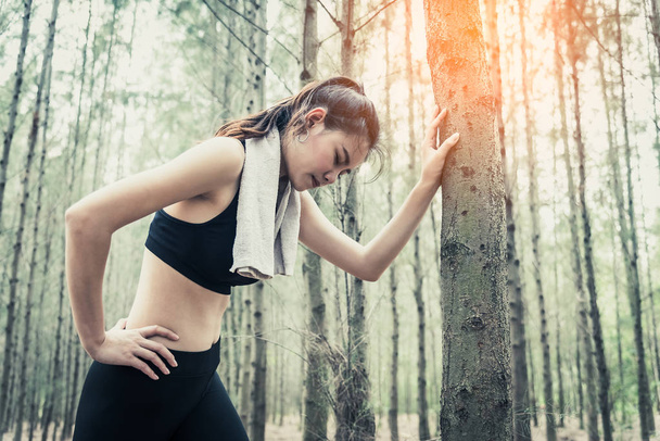 Asian beauty woman tiring from jogging in forest. Lean on tree. Towel and sweat elements. Sport and Healthy concept. Jogging and Running concept. Relax and waist pain theme. Outdoors activity theme. - Photo, Image