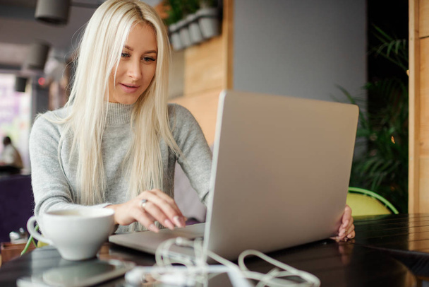 Mujer joven de confianza en ropa casual trabajando en el ordenador portátil mientras está sentado en la oficina creativa o cafetería y woking. Sosteniendo una taza de café y sonriendo
  - Foto, imagen