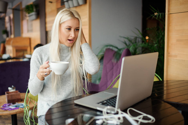 Shocked young woman in casual wear working on laptop while sitting in creative office or cafe and woking. Holding a cup of coffee and smiling  - Φωτογραφία, εικόνα