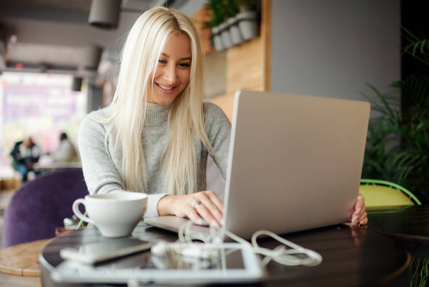Confident young woman in casual wear working on laptop while sitting in creative office or cafe and woking. Holding a cup of coffee and smiling  - Foto, imagen