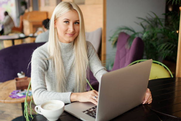 Confident young woman in casual wear working on laptop while sitting in creative office or cafe and woking. Holding a cup of coffee and smiling  - Zdjęcie, obraz