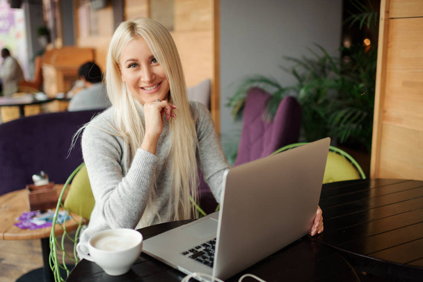 Confident young woman in casual wear working on laptop while sitting in creative office or cafe and woking. Holding a cup of coffee and smiling  - Fotografie, Obrázek