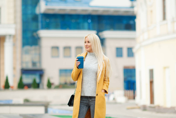 外出先でコーヒー。コーヒー カップを押しながら通りを歩いて笑顔明るい黄色のコートで若い金髪美女  - 写真・画像