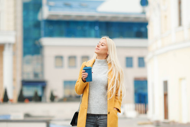 Coffee on the go. Beautiful young blond woman in bright yellow coat holding coffee cup and smiling while walking along the street  - Foto, Bild