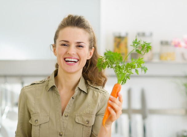 Smiling young housewife holding carrot in kitchen - Valokuva, kuva