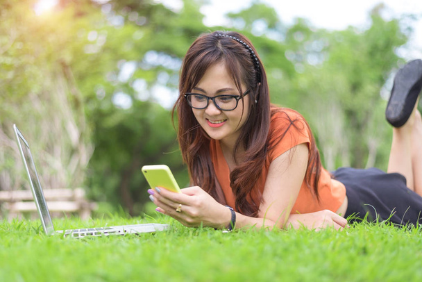 Happy Asian woman using smartphone when relaxing in park. People and lifestyle concept. Technology and Relaxation theme. Outdoors leisure and activity theme. Laptop computer element - Foto, Bild