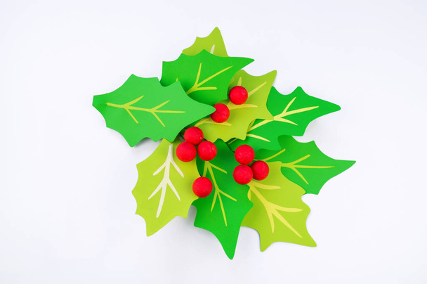 Flower holly made of paper crafts. Red berry from the ball. Christmas leaves. White background. Children's creativity. - Foto, Imagen
