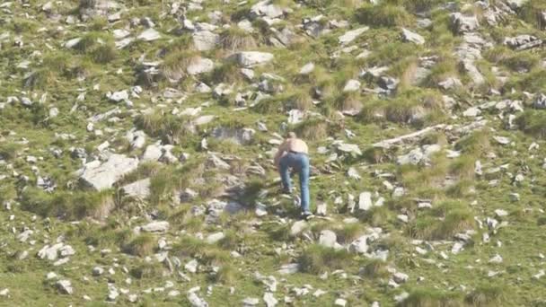 Geri taş tepe tırmanma adamdan görünümünü. Hisse senedi. Erkek kot pantolon ve gömleksiz taş tepenin yamaçlarında yeşil çim ile yukarı tırmanıyor - Video, Çekim