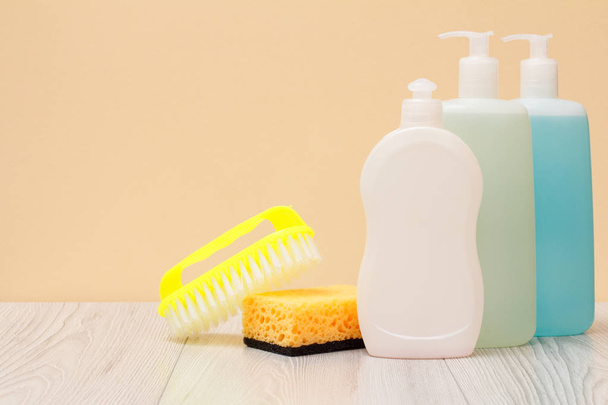 Plastic bottles of dishwashing liquid, glass and tile cleaner, brush and sponge on wooden boards and beige background. Washing and cleaning concept. - Foto, immagini