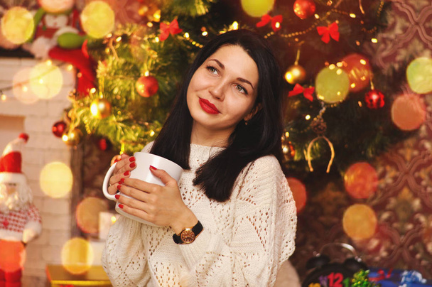 Una hermosa joven se sienta en un suéter blanco con una gran taza de bebida caliente bajo el árbol de Navidad y sueña
. - Foto, imagen