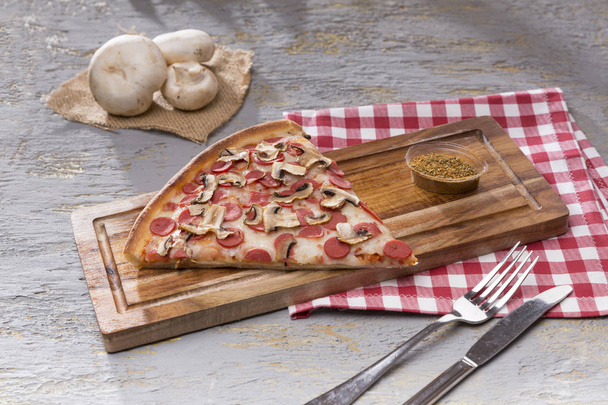 Nahaufnahme eines Stücks italienische Pizza und Gewürze auf einem Holzschneidebrett vor grauem Hintergrund - Foto, Bild