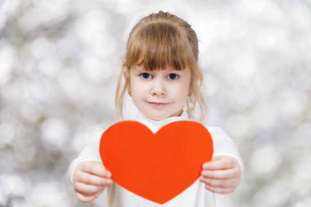 A tender smiling child is showing a big bright red heart in her hands at the light blurred background. The concept is the children's rights defending principle. - Foto, Imagen