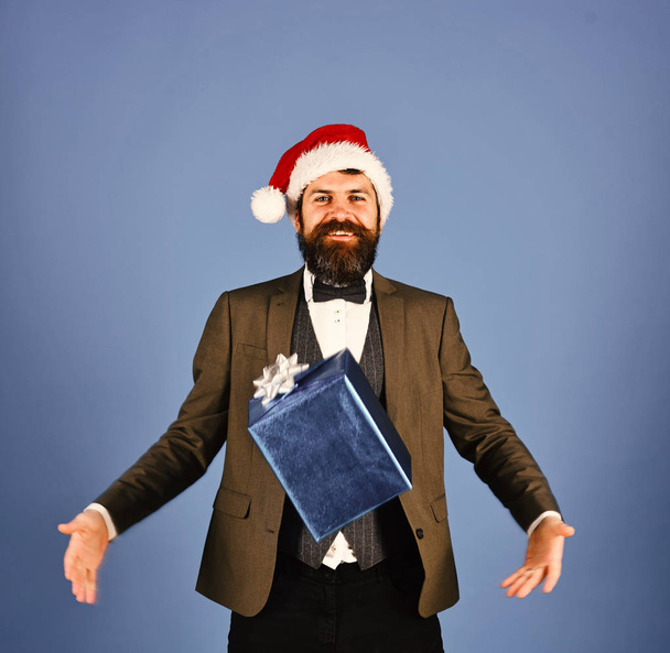 Businessman with happy face catches blue present box. Manager with beard and flying Christmas gift. Man in smart suit and Santa hat on blue background. Business and celebration concept. - Fotografie, Obrázek