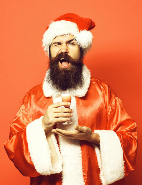 handsome bearded santa claus man with long beard on smilingface holding glass of alcoholic shot on hand in christmas or xmas sweater and new year hat on red studio background - Foto, Imagem